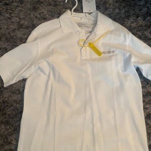 Vit piké från Les Deux - Fräsch vit piké från Les Deux med klassisk krage och korta ärmar. T-shirten har broderad logga på bröstet och knappar vid halsen. Materialet är mjuk bomull som känns skönt mot huden. Perfekt för dig som gillar stilren och enkel design.