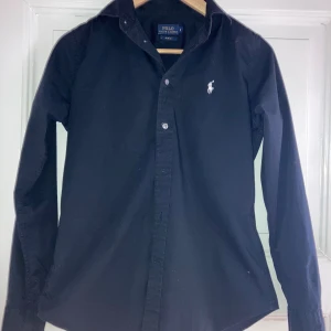 Svart skjorta från Polo Ralph Lauren - Svart skjorta från Polo Ralph Lauren med klassisk krage och vit broderad logga på bröstet. Slim fit-modell med långa ärmar och knappar framtill. Tillverkad i mjuk bomull som känns skön mot huden. Perfekt för dig som gillar stilrena och tidlösa plagg.