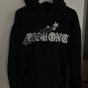 Svart hoodie från Demont med stjärnor - Svart hoodie från Demont i storlek L med stor vit broderad logga och coola stjärndetaljer på bröstet. Tröjan har huva och klassisk känguruficka framtill. Perfekt för dig som gillar streetwear och vill sticka ut med snygga detaljer. Väldigt bra skick