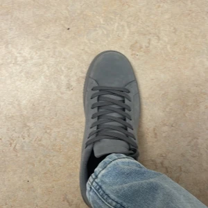 Grå skor  - Snygga grå sneakers  med stilren design och platt sula. Skorna har rund tå, snörning och är tillverkade i mocka-liknande syntetmaterial. Perfekt för dig som gillar minimalistisk look och vill ha ett par neutrala sneakers till jeans eller streetwear.
