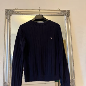 Mörkblå ribbstickad tröja från GANT - Mörkblå ribbstickad tröja från GANT i klassisk modell med rund hals och diskret logga på bröstet. Tröjan har lång ärm och är tillverkad i mjuk bomull, perfekt för lager-på-lager och chill stil.