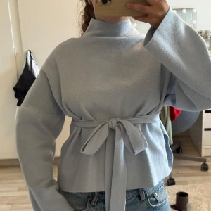 Stickad tröja med öppen rygg - Supersnygg ljusblå stickad tröja med hög krage och breda ärmar. Tröjan har ett knytband i midjan och en öppen rygg med cool twistdetalj. 