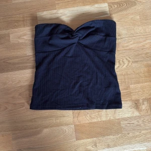 Svart bandeau tubtopp från Stockh LM Studio - Svart bandeau tubtopp från Stockh LM Studio med snygg knutdetalj framtill. Toppen är ribbad och axelbandslös, perfekt för en trendig look. Materialet är stretchigt och sitter tight mot kroppen.