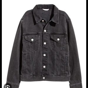 Svart jeansjacka med vita knappar - Snygg svart jeansjacka med klassisk krage och silver knappar framtill. Jackan har två bröstfickor med lock och raka sömmar som ger en clean look. Perfekt för dig som gillar en stilren och tidlös vibe.