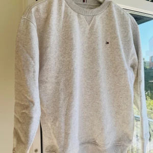 Ljusgrå sweatshirt från Tommy Hilfiger - Säljer en stilren ljusgrå sweatshirt från Tommy Hilfiger med diskret logga broderad på bröstet. Tröjan har rund hals, ribbade muddar och är långärmad. Perfekt för dig som gillar klassisk och enkel design med en touch av street. Knappt använd. Mycket gott skick.