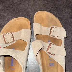 Beige mockasandaler från Birkenstock - Snygga sandaler från Birkenstock i beige mocka med två breda remmar och justerbara metallspännen. Fotbädden är klassiskt formad och sulan har ett vågigt mönster för extra grepp. Perfekta för chill dagar och enkel stil.