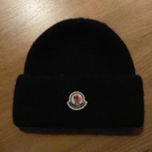 Svart Moncler mössa i ull - Svart ribbstickad mössa från Moncler med klassisk uppvikt kant och broderad Moncler-logga framtill. Tillverkad i mjuk ull som håller dig varm och ger en clean look. Perfekt accessoar för kyliga dagar och snygg till streetwear.