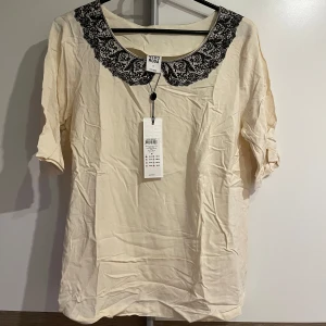 Beige blus med spetskrage Vero Moda - Supersöt beige blus från Vero Moda med svart spetsdetalj runt kragen. Blusen har halvkort ärm och en lös passform som ger en chill vibe. Perfekt för dig som gillar romantiska detaljer och vill sticka ut lite extra.