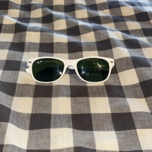 Vita Ray-Ban Wayfarer solglasögon - Snygga vita Ray-Ban Wayfarer solglasögon med klassisk form och mörkgröna glas. Ikonisk logga på sidan och cool retrostil som lyfter vilken outfit som helst. Perfekta för dig som vill sticka ut med stilren design och tidlös vibe.