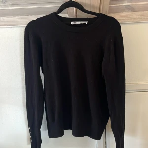 Svart långärmad tröja från Zara - Svart långärmad tröja från Zara i ribbad bomullsmix. Klassisk rund halsringning och smal passform. Ärmarna har dekorativa knappar vid ärmslutet. Perfekt basic-plagg att styla med jeans eller kjol. Köpte den ifrån Sellpy och därmed va den sönder i ärmarna redan från när jag köpte tröjan!