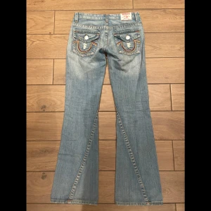 True Religion ljusblå bootcut jeans - Säljer ett par ljusblå jeans från True Religion med bootcut passform. Jeansen har slitna detaljer framtill, klassiska kontrastsömmar och stora fickor bak med broderade detaljer och knappar. Tillverkade i jeansmaterial med låg midja.