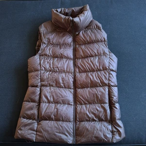 Brun dunväst med hög krage - Snygg brun dunväst med quiltad design och hög krage. Västen har dragkedja framtill och fikor på båda sidorna och är perfekt att slänga över en hoodie eller tröja för extra värme. Enkel och stilren look som funkar till många outfits.