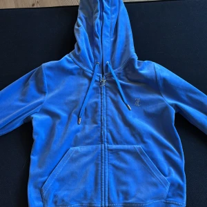 Blå Juicy Couture velour hoodie och byxa - Blå hoodie från Juicy Couture i mjuk velour med huva, snörning och dragkedja. Stora fickor framtill och ikonisk Juicy-text i strass på ryggen. Blå mjukisbyxa från Juicy Couture i mjuk velour med snörning och fickor fram och baktill. Man kan köpa tröjan och byxorna enskilt för 800kr styck.