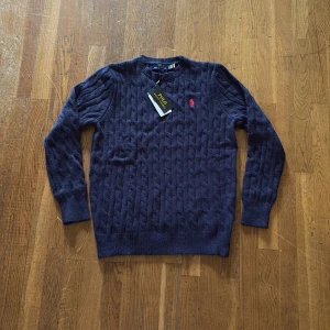 Mörkblå stickad tröja Polo Ralph Lauren - Snygg mörkblå stickad tröja från Polo Ralph Lauren med klassiskt kabelstickat mönster och röd broderad logga på bröstet. Rund hals och långa ärmar, perfekt för dig som gillar stilrena och tidlösa plagg. Passar dig som vill ha en chill men ändå preppy look.