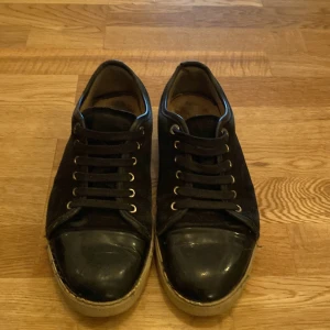 Svarta sneakers från Lanvin i mocka och skinn - Snygga svarta sneakers från Lanvin med ovandel i mocka och tåparti i blankt skinn. Skorna har svarta snören, rund tå och vit sula med logga undertill. Tillverkade i Portugal och har en stilren, klassisk look som passar till det mesta.