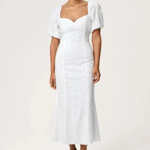 Adoore klänning vit VERCELLI modellen - Supersnygg vit långklänning med puffärmar och v-ringad hals. Klänningen har ett subtilt blommigt mönster, öppen rygg med knyt och slits baktill. Passformen är slim och materialet känns som linne. Perfekt för många tillfällen! Använd endast en gång❤️