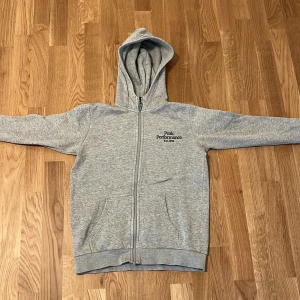 Grå hoodie från Peak Performance - Snygg grå hoodie från Peak Performance med dragkedja och huva. Broderad logga på bröstet och två fickor framtill. Perfekt för chill dagar eller när du vill ha en enkel och stilren look.