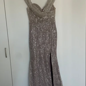 Champagnefärgad glittrig långklänning - Säljer en elegant långklänning i champagnefärg med glittriga paljetter över hela klänningen. Modellen har omlott-detalj vid axlarna, v-ringad urringning och en snygg slits framtill. Perfekt för bal eller fest! Materialet är syntet med paljetter & pärlor för extra glam. Den är lite tyngd med ett litet släpp. 