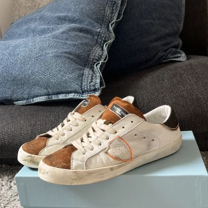 Philippe Model sneakers i skinn och mocka - Snygga Philippe Model sneakers med vit skinnbas, bruna mockadetaljer på tå och plös samt svart hälparti. Klassisk låg modell med snörning och rund tå. Orange söm runt skölden på sidan ger en cool touch. Perfekta för dig som gillar streetstyle. 36 fast passar 37