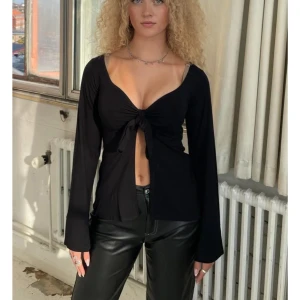 Svart långärmad knytblus från design by si - Svart långärmad topp med djup v-ringning och knyt framtill. Modellen är croppad och har en snygg, figurnära passform. Perfekt för dig som gillar trendiga och stilrena plagg med en twist.❤️