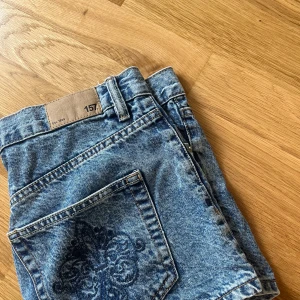 Blå jeansshorts från 157 - Snygga blå jeansshorts från 157 med klassisk femficksdesign och broderad detalj på bakfickan. Shortsen har hög midja, bälteshällor och knappar framtill. 