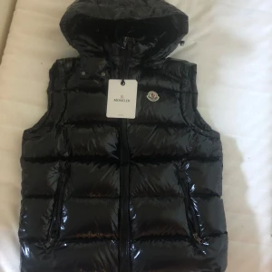 Svart dunväst från Moncler med huva - Säljer en glansig svart dunväst från Moncler med avtagbar huva och dragkedja framtill. Västen har två fickor med dragkedja och ikonisk Moncler-logga på bröstet. Insidan har ett coolt serietryck och Moncler-märke. Perfekt för lager-på-lager-stil.