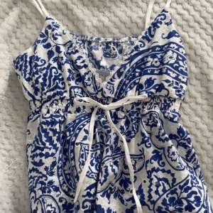 Vit och blå babydoll topp - Säljer en söt babydoll topp med tunna axelband och v-ringning. Klänningen är vit med blått blommigt mönster och har en snörning i midjan för extra passform. Materialet känns lätt och luftigt, perfekt för varma dagar. Spetsdetaljer vid halsringningen ger en extra touch.