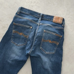  Nudie Jeans - Snygga blå jeans från Nudie Jeans med klassisk femficksdesign och orange sömmar på bakfickorna. Jeansen har en smilfit passform och är tillverkade i slitstarkt bomullstyg. Perfekta för dig som gillar en tidlös och avslappnad stil.modell grim Tim 