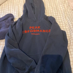 Mörkblå hoodie Peak Performance - Mörkblå hoodie från Peak Performance med röd broderad logga på bröstet. Klassisk huva och känguruficka framtill. Perfekt för dig som gillar en enkel och stilren look med sportig vibe.