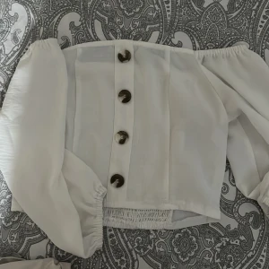 Vit offshoulder blus med knappar - Trendig vit blus med offshoulder-modell och långa puffärmar. Framtill sitter stora, snygga knappar i mörkare ton som ger en cool kontrast. Smockad resår bak för skön passform. Perfekt för dig som vill sticka ut med en stilren look.