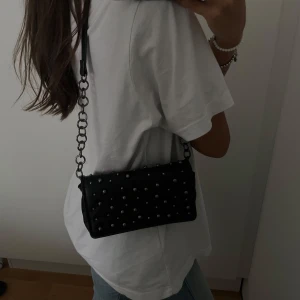 Svart axelväska med silvriga runda nitar och kedja - En väldigt unik och svart axelväska med silverfärgade runda nitar. Väskan har en rektangulär form och är tillverkad i skinnimitation. Man kan ha kedjan både lång och kort beroende på vad man gillar. Väskan passar superbra till alla outfits 🖤