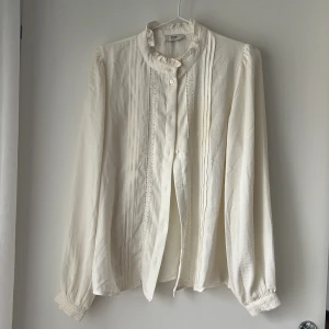 Vit blus med volangkrage från JDY - Supersöt vit blus från JDY med volangkrage och knappar framtill. Långa ärmar med spetsdetaljer vid ärmslut och längs knappslån. Luftigt material och klassisk passform, perfekt för en stilren look.