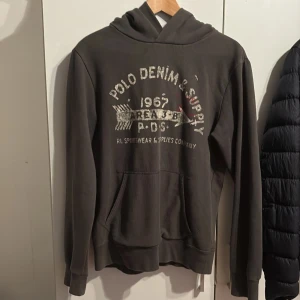 Mörkgrå hoodie från Ralph Lauren - Säljer en snygg mörkgrå hoodie från Ralph Lauren, Denim & Supply. Den har ett coolt tryck på framsidan med texten 'Polo Denim & Supply 1967'. Perfekt för en avslappnad stil och passar bra till jeans. Den är i bra skick och superbekväm att ha på sig. Perfekt för höst och vår!