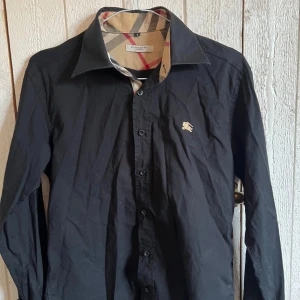 Svart Burberry skjorta  - Svart skjorta från Burberry med klassiska rutiga detaljer på insidan av kragen och ärmsluten. Jättesnygg och stilren. Storlek S, men något liten i storleken. Säljer för att jag inte fått någon användning av den. Den är något skrynklig på bilderna, men jag kan stryka den innan jag skickar iväg den. ☺️