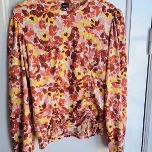 Blommig långärmad topp från Gina Tricot - Superfin långärmad topp från Gina Tricot med blommigt mönster i rött, gult och vitt. Toppen har puffiga axlar och är tillverkad i ett lätt, ribbat material som ger en snygg struktur. Perfekt för dig som vill sticka ut med färg och mönster!