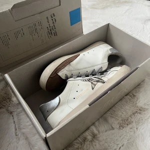 Golden Goose Superstar sneakers - Golden Goose Superstar sneakers i vitt skinn med silverfärgade detaljer på hälen, grå snörning och bruna mockadetaljer på tån. Skorna har en zebra stjärna på sidan. Använt skick men inga hål eller liknande. Lådan tillkommer, skriv för fler bilder! Pris kan diskuteras