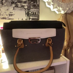 River Island grå handväska med kedja - Snygg handväska från River Island i grå filt med svarta och vita detaljer. Väskan har ljusbruna handtag i skinnimitation, guldfärgad kedja och dragkedja upptill. Perfekt storlek för allt du behöver och stilren design med metallogo framtill.