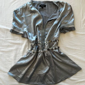 Silvrig blus med knappar från Kriss - Supersnygg blå-silvrig blus från Kriss i glansigt satinliknande material. Modellen har korta ärmar, markerad midja med dubbla rader av dekorativa knappar i olika färger och v-ringad hals. Perfekt för dig som vill sticka ut med en unik detalj! 💞