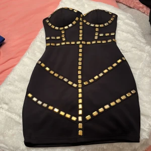 Svart kort klänning med gulddetaljer - Säljer en svart axelbandslös kort klänning med snygga guldfärgade dekorstenar framtill och en dragkedja i ryggen. Klänningen har en figurnära passform och markerad byst, perfekt för dig som vill sticka ut med coola detaljer och en modern look.