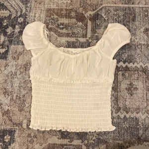 Vit blus med puffärmar och smock - Supersöt vit blus med puffiga korta ärmar och smockad midja. Toppen har en rynkad, luftig överdel och små volangdetaljer vid nederkanten. Perfekt för dig som gillar romantisk och feminin stil.