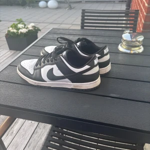 Nike Dunk Low svart/vit sneakers - Säljer ett par Nike Dunk Low sneakers i klassisk svart och vit färg. Skorna har svart swoosh, svarta snören och vit tåbox med perforeringar. Låg modell med platt sula och rund tå. Perfekta för dig som gillar streetstyle och vill ha en ikonisk look.