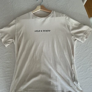 Beige t-shirt från Lyle & Scott Junior - Beige t-shirt från Lyle & Scott Junior med svart broderad logga på bröstet och gul logga på ärmen. Klassisk rund hals och korta ärmar. Tillverkad i mjuk bomull för en skön känsla och enkel stil.