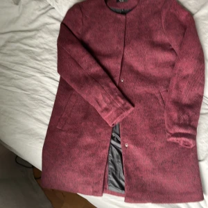 Vinröd kappa från ONLY, storlek S - Säljer en vinröd kappa från ONLY i storlek S. Jackan har en rak och stilren modell med rund halsringning, knäppning framtill och diskreta fickor på sidorna. Materialet är ullblandat och fodret är svart. Perfekt för höst och vinter!