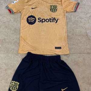 FC Barcelona Lewandowski bortatröja set - FC Barcelona bortaställ med Lewandowski 9. Tröjan är guldbeige med kartmönster, svarta detaljer och klubbmärke, samt Spotify-logga. Korta ärmar med färgglada muddar. Matchande mörkblå shorts med klubbmärke och Nike-logga. Materialet är lätt och andas.