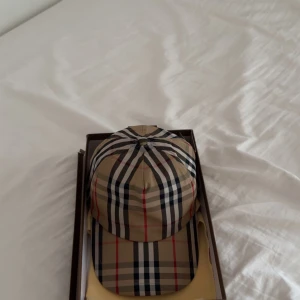 Beige rutig keps från Burberry - Snygg keps från Burberry i klassiskt beige rutigt mönster med inslag av svart, vitt och rött. Kepsen har böjd skärm och justerbar rem bak. Perfekt accessoar för att lyfta din outfit med en lyxig touch.