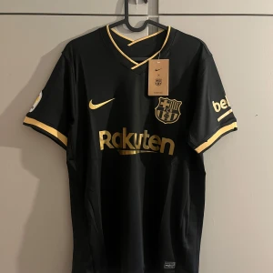 Svart Barcelona fotbollströja Nike S - Barcelona fotbollströja i svart med guldiga detaljer och tryck från Nike. Tröjan har klubbmärke, sponsorloggor och La Liga-märke på ärmen. V-ringad hals med guldkant och korta ärmar. Materialet är lätt och andas, perfekt för match eller träning.