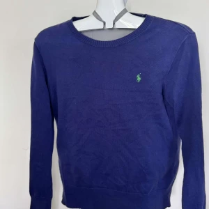 Blå tröja från Polo Ralph Lauren - Mörkblå tröja från Polo Ralph Lauren med rund halsringning och klassisk grön logga broderad på bröstet. Tröjan är långärmad och tillverkad i mjuk bomull, perfekt för en stilren och enkel look.