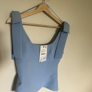 Ljusblå linnetopp med rosetter - Superfin ljusblå topp  från Zara med breda axelband som knyts till stora rosetter på axlarna. Toppen har en figurnära passform och är tillverkad i ett slätt, mjukt material. Perfekt för att ge en romantisk och somrig touch till din outfit.