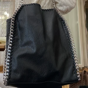 Svart axelväska med kedjedetaljer Tiamo - Svart axelväska från Tiamo i skinnimitation med silverfärgade kedjor längs kanterna och som axelrem. Väskan har en dragkedjeficka på baksidan och flera fack inuti. Endast använd ett fåtal gånger. 
