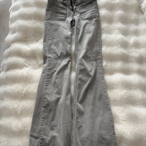 Grå bootcut jeans från Nelly - Snygga grå jeans från Nelly med bootcut-modell och hög midja. Jeansen har breda ben, framfickor och bakfickor med lock och knapp. Tillverkade i mjuk bomullsdenim med dragkedja och dubbla knappar framtill.